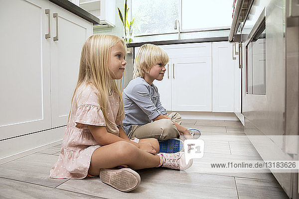 Brother and sister sitting on the floor in kitchen looking at oven