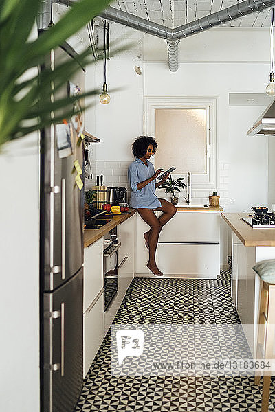 Frau sitzt auf der Arbeitsplatte ihrer Küche und benutzt morgens ein digitales Tablett