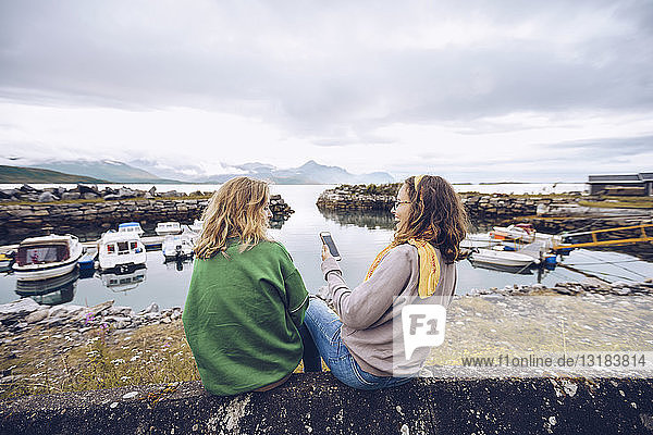 Norwegen  Senja  zwei junge Frauen sitzen mit einem Mobiltelefon an einem kleinen Hafen
