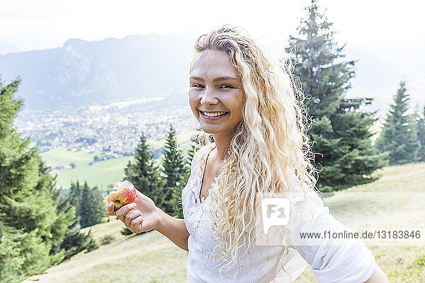 Deutschland  Bayern  Oberammergau  Porträt einer lächelnden jungen Frau  die auf einer Bergwiese einen Apfel isst