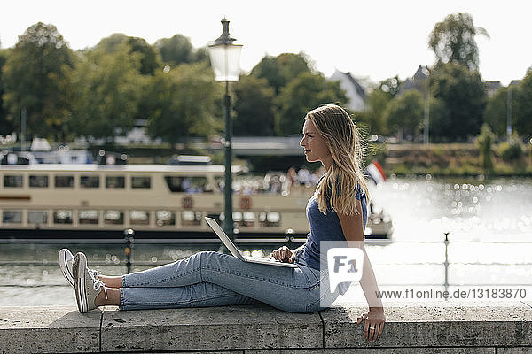 Niederlande  Maastricht  junge Frau sitzt mit Laptop auf einer Mauer am Flussufer