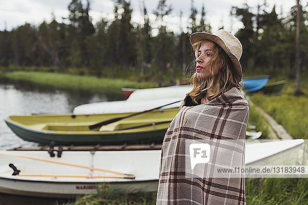 Finland  Lapland  woman wearing a hat wrapped in a blanket standing at the lakeside