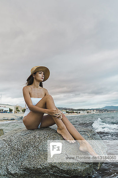 Schöne junge Frau im Badeanzug und mit Hut auf einem Felsen im Meer sitzend