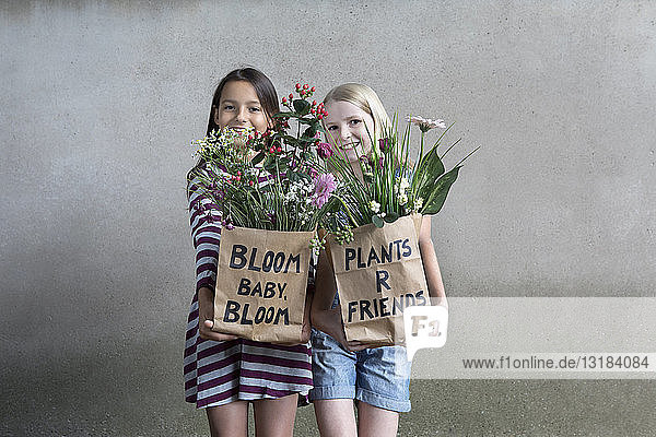 Porträt von zwei lächelnden Mädchen  die nebeneinander stehen und Papiertüten mit Blumen anbieten