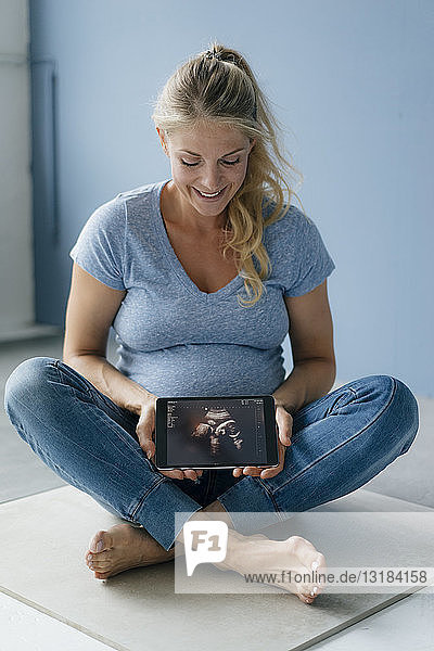 Lächelnde schwangere Frau sitzt auf dem Boden und zeigt Ultraschallbild auf Tablette