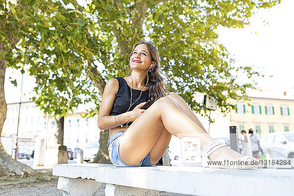 Lächelnde junge Frau entspannt sich auf einer Parkbank und hört Musik