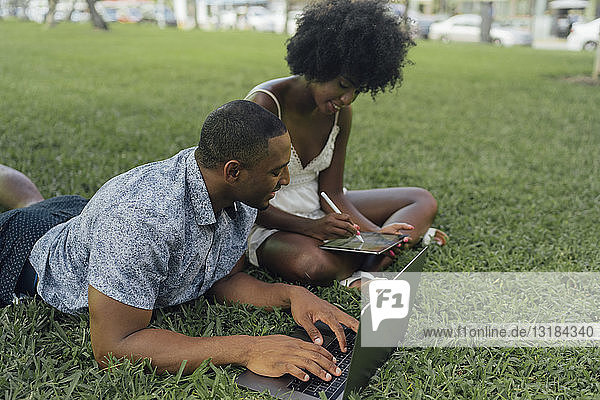 Young couple using tablet and laptop on lawn in a park