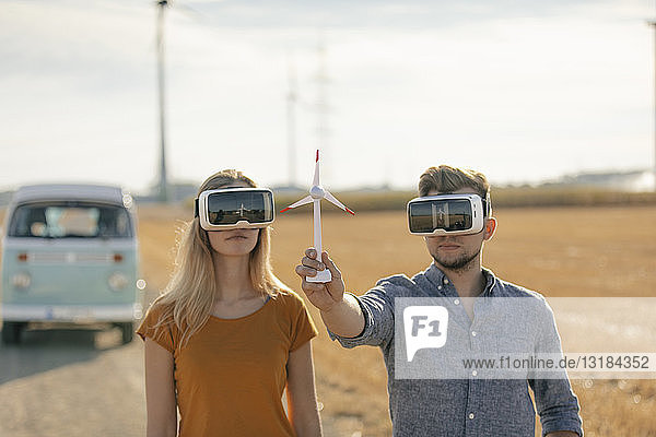 Junges Paar mit VR-Brille am Wohnmobil in ländlicher Landschaft hält Modell einer Windkraftanlage