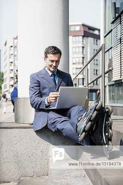 Businessman with inlineskates sitting down using laptop in the city