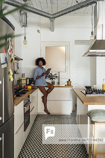 Frau sitzt auf der Arbeitsplatte ihrer Küche und benutzt morgens ein digitales Tablett