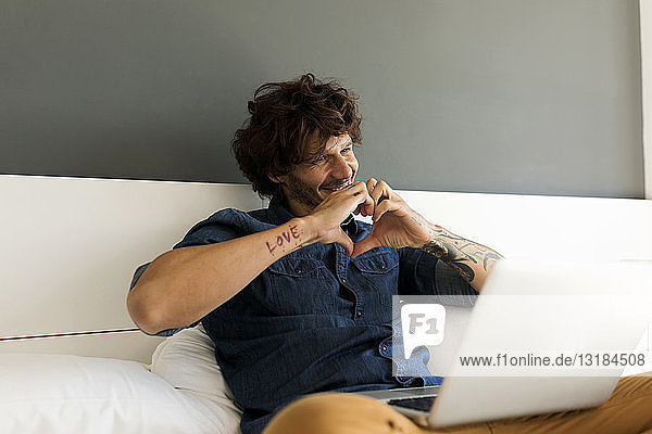 Glücklicher tätowierter Mann mit Laptop chattet mit seiner Freundin im Bett
