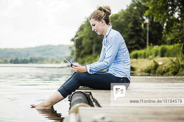 Frau sitzt mit Tablette auf einem Steg an einem See