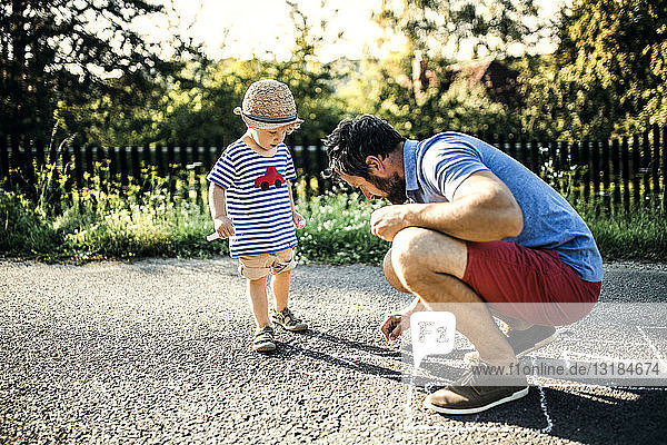 Vater zeichnet Hopscotch auf Asphalt  während sein kleiner Sohn ihm zusieht