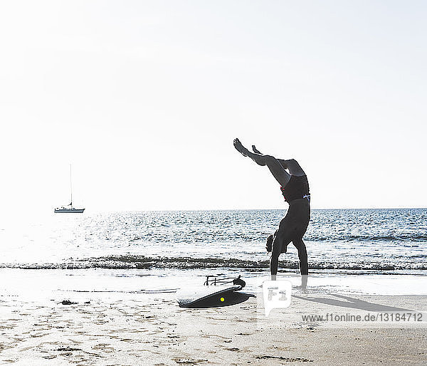 Frankreich  Bretagne  junger Mann macht einen Handstand am Strand neben dem Surfbrett