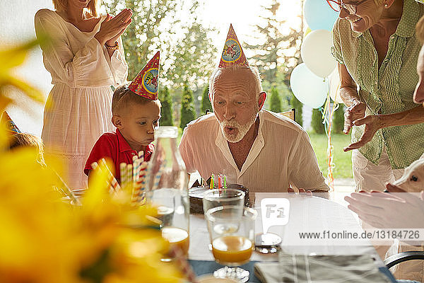 Glückliche Großfamilie beim Kerzenausblasen der Geburtstagstorte auf einer Gartenparty
