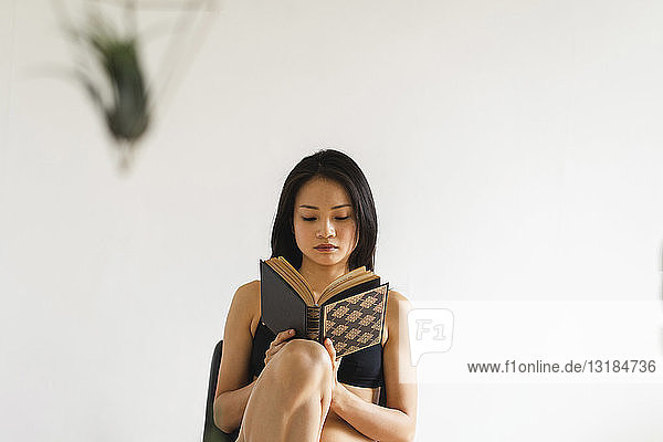 Attraktive junge Frau in Dessous sitzt zu Hause auf einem Stuhl und liest ein Buch
