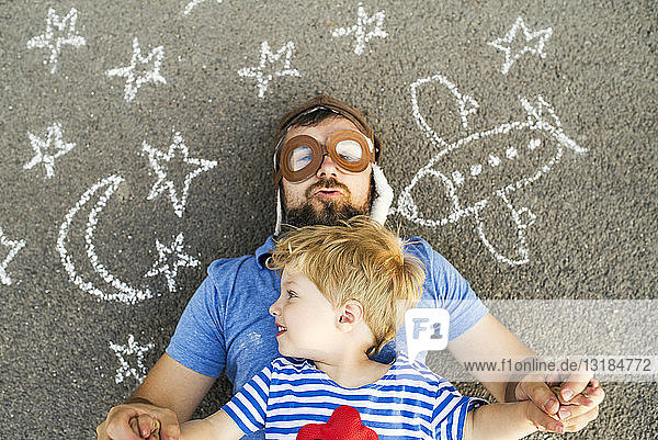 Porträt eines reifen Mannes mit Pilotenhut und seinem kleinen Sohn auf Asphalt liegend  gemalt mit Flugzeug  Mond und Sternen