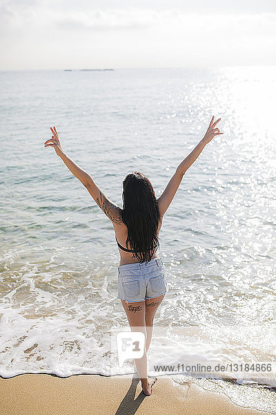 Rückenansicht einer jungen Frau  die am Meeresufer steht und ein Siegeszeichen zeigt