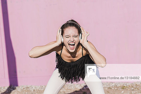 Überschwängliche junge Frau mit Kopfhörern schreit vor rosa Wand
