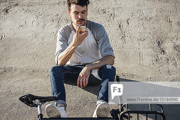 Junger Mann mit Pendler-Fixie-Fahrrad sitzt mit Handy an Betonwand