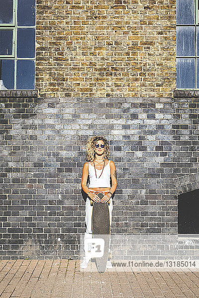 Junge Frau steht mit Skateboard an Ziegelmauer in der Stadt