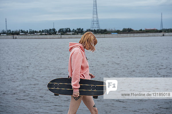 Junge Frau hält Carver Skateboard in der Hand und geht am Flussufer