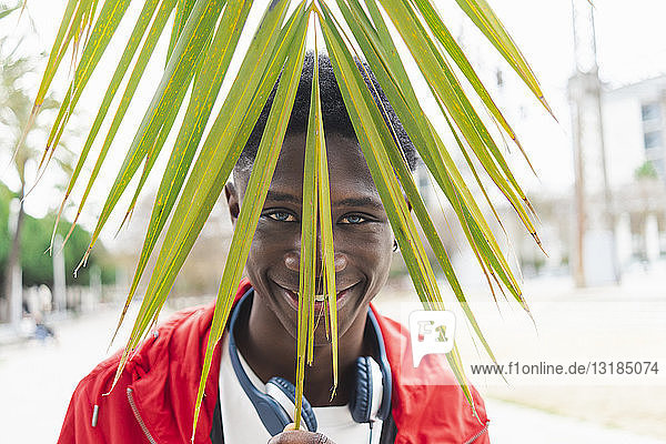 Junger schwarzer Mann mit Kopfhörern hinter einem Palmenblatt  lächelnd  Porträt