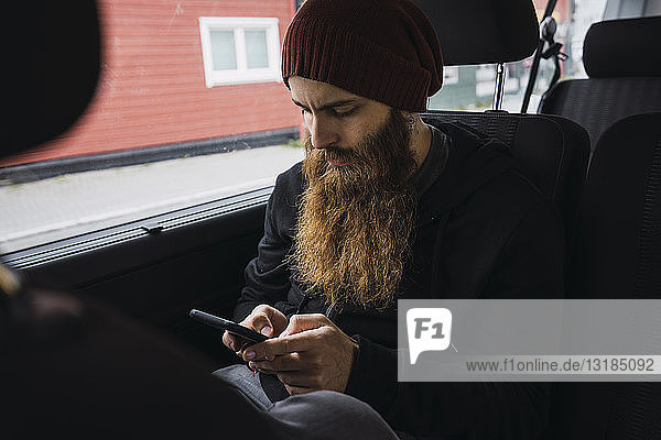Bärtiger junger Mann sitzt auf dem Rücksitz eines Autos und überprüft sein Handy