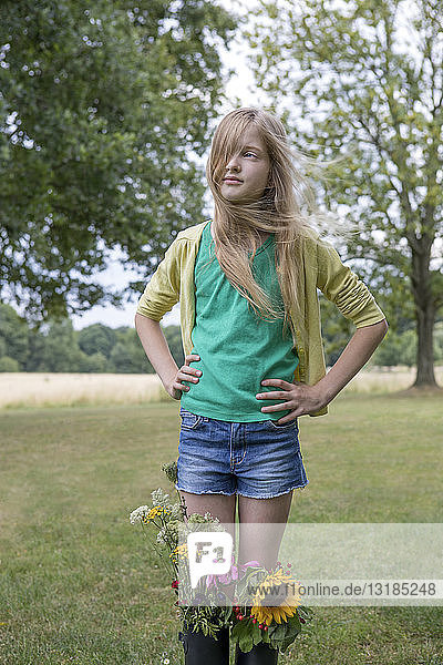 Portrait of blond girl standing on a meadow with bunches of flowers in her rubber boots