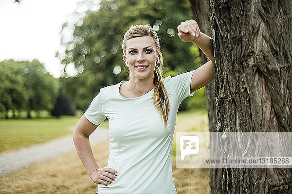 Lächelnde  sportliche junge Frau  die sich an einen Baum in einem Park lehnt