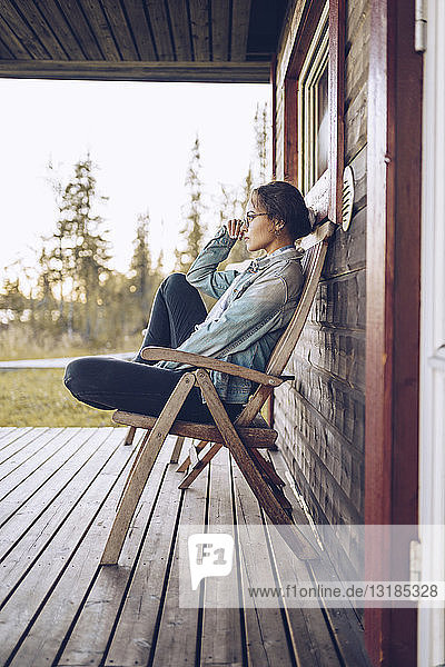 Schweden  Lappland  nachdenkliche junge Frau sitzt auf einem Stuhl auf der Veranda und schaut in die Ferne