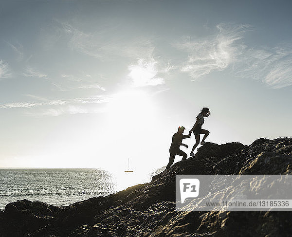 Frankreich,  Bretagne,  junges Paar klettert bei Sonnenuntergang auf einen Felsen am Strand
