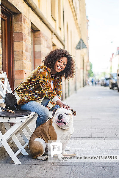 Smiling woman sitting with dog on sidewalk in city