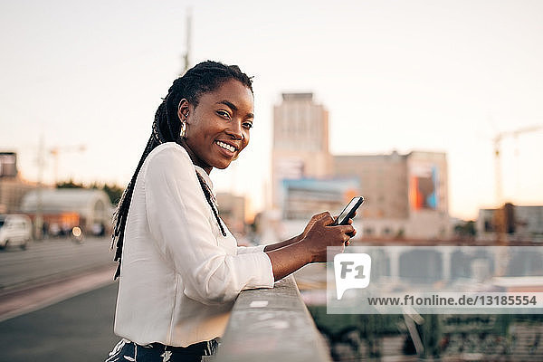 Side view portrait of young woman holding mobile phone while standing on bridge in city