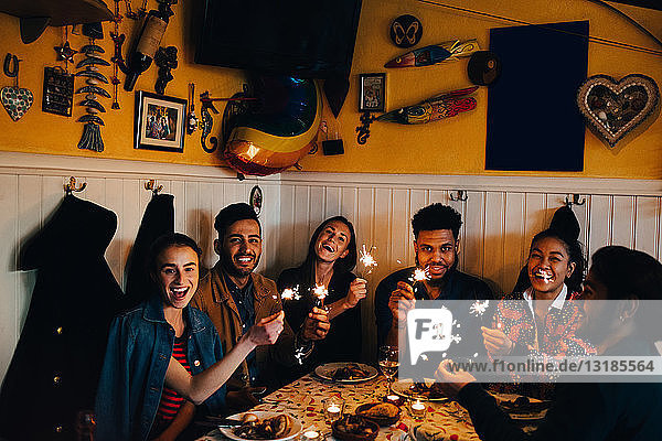 Porträt fröhlicher junger multi-ethnischer Freunde  die mit brennenden Wunderkerzen am Tisch im Restaurant sitzen