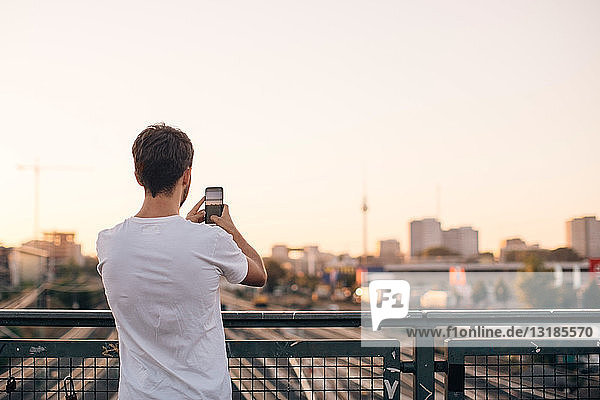 Rückansicht eines jungen Mannes  der die Stadt mit seinem Mobiltelefon fotografiert  während er auf der Brücke vor klarem Himmel steht
