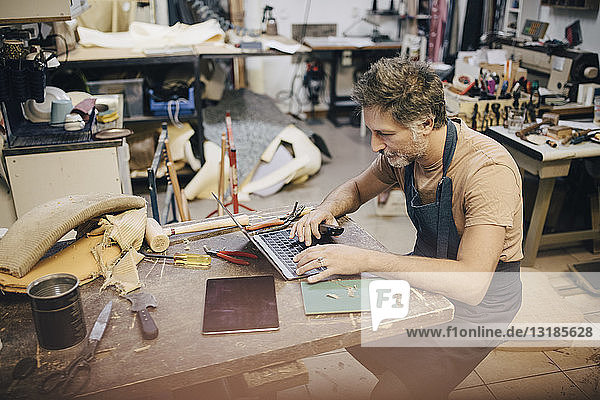 Male craftsperson using laptop at workbench in workshop