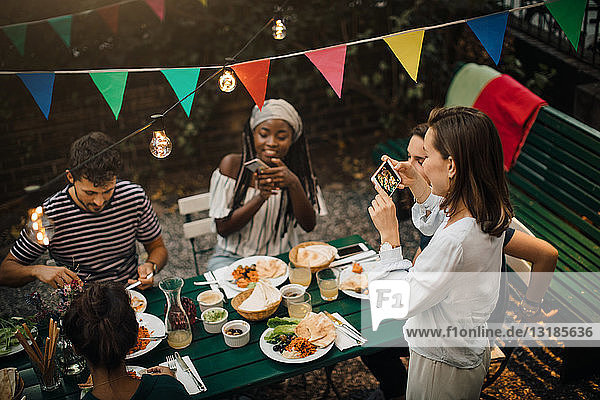 High angle view of young women photographing friends having food during dinner party in backyard