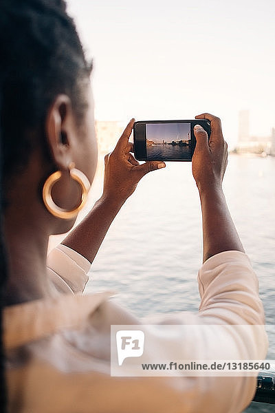 Cropped image of young woman photographing river through smart phone in city
