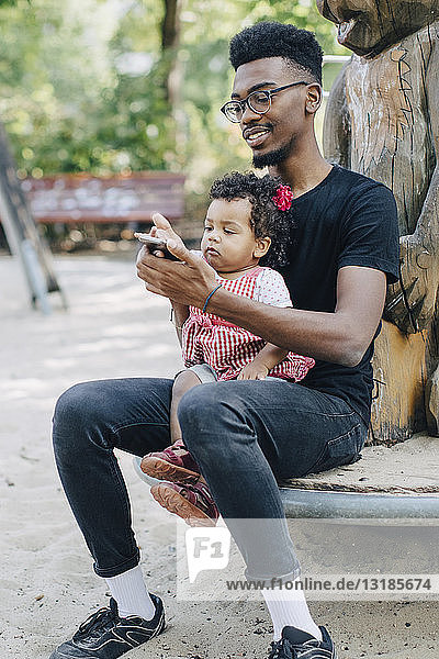 Kleinkind benutzt Mobiltelefon  während es mit seinem Vater auf einem Spielgerät im Freien auf dem Spielplatz sitzt