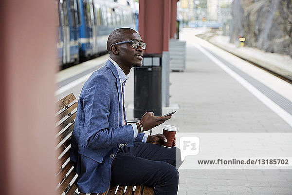 Thoughtful businessman sitting with smart phone and coffee at railroad station