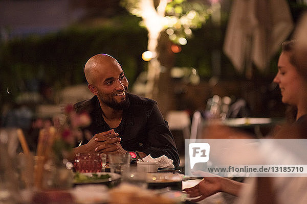 Lächelnder junger Mann mit rasiertem Kopf schaut eine Freundin an  während er während der Dinnerparty am Tisch sitzt