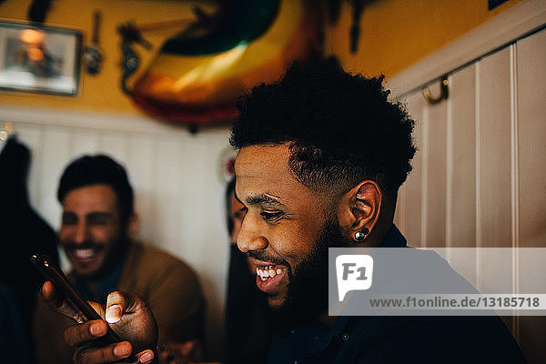 Lächelnder junger Mann benutzt Smartphone  während er im Restaurant bei multiethnischen Freunden sitzt
