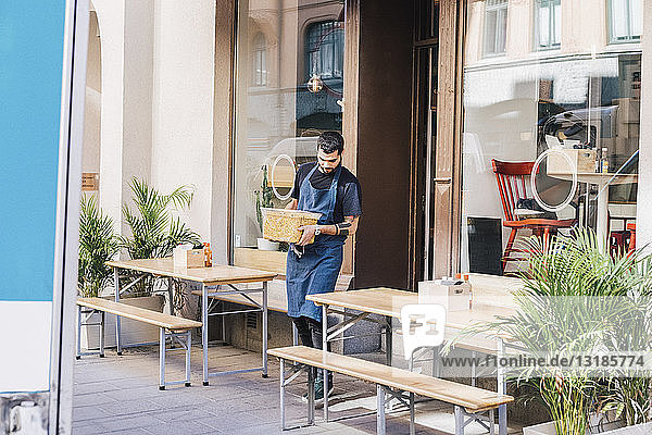 Junger männlicher Besitzer trägt in voller Länge einen Lebensmittelbehälter inmitten von Möbeln gegen ein Restaurant