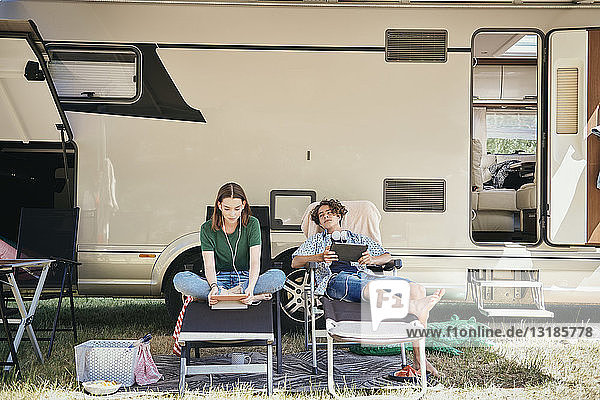 Teenager-Geschwister sehen sich einen Film auf einem digitalen Tablet an  während sie auf Klappstühlen gegen ein Wohnmobil auf dem Campingplatz sitzen