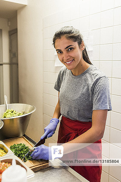 Lächelnder junger Chefkoch beim Zerkleinern von Gemüse auf dem Schneidebrett in der Großküche eines Restaurants