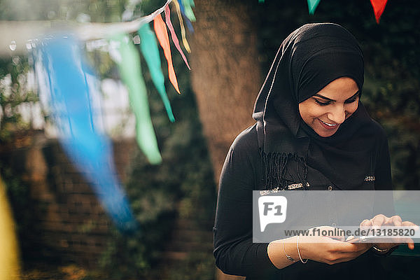 Lächelnde junge Frau im Hidschab mit Mobiltelefon im Hinterhof