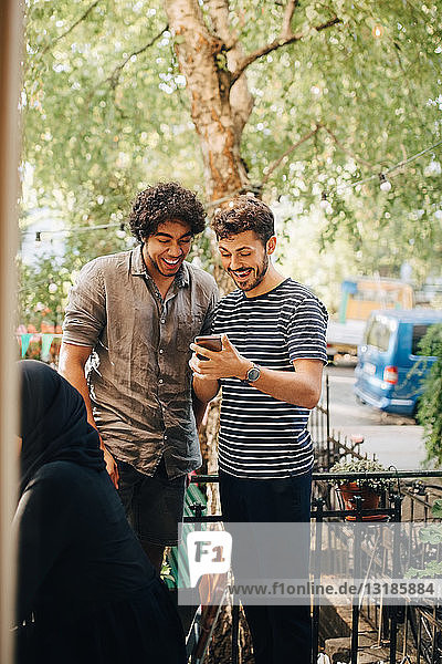 Fröhlicher junger Mann zeigt einem männlichen Freund ein Smartphone  während er während der Party auf dem Balkon steht