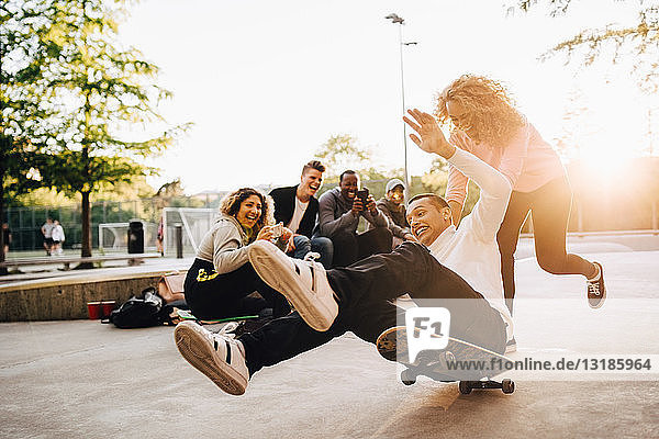 Lachende Freunde fotografieren Mann  der vom Skateboard fällt  während Frau ihn im Park schubst