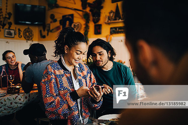 Lächelnde junge Frau teilt Smartphone mit männlichem Freund  während sie während der Dinnerparty im Restaurant sitzt